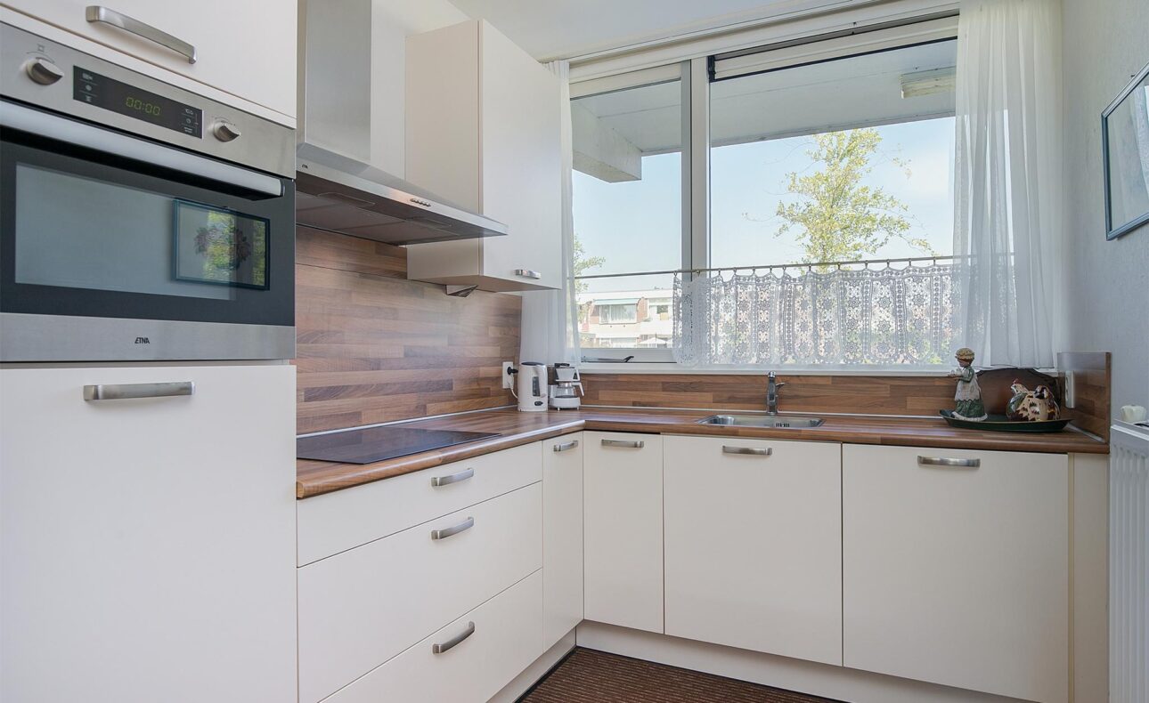 Moderne keuken met witte kasten, houten werkblad, ingebouwde oven en kookplaat. Groot raam met uitzicht en gordijn.