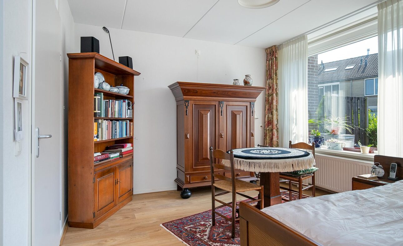 Woonkamer met houten meubels, een boekenkast, een ronde tafel met kleed, en een groot raam met uitzicht op de tuin.