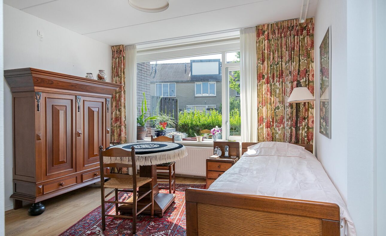 Een gezellige kamer met een houten kast, een eenpersoonsbed, een ronde tafel met stoelen en een groot raam met uitzicht op de tuin.