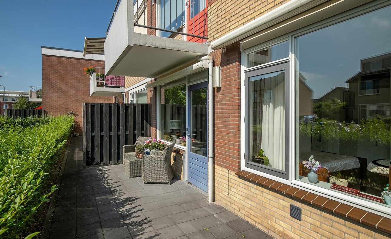 Buitenzijde van een modern bakstenen gebouw met grote ramen en een betegeld terras, omgeven door groene heggen onder een blauwe lucht.