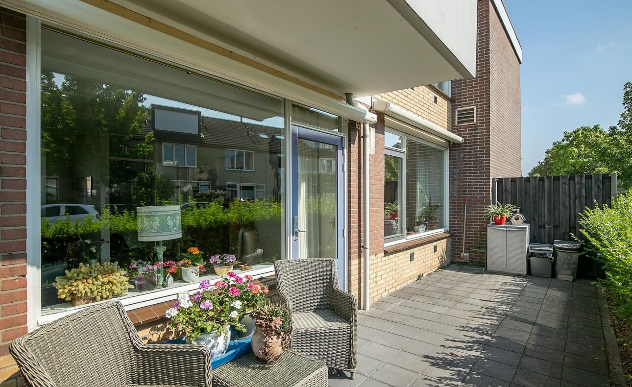 Twee rotan stoelen met kussens en een tafel met bloemen op een betegeld terras voor een huis met grote ramen en groene heg.