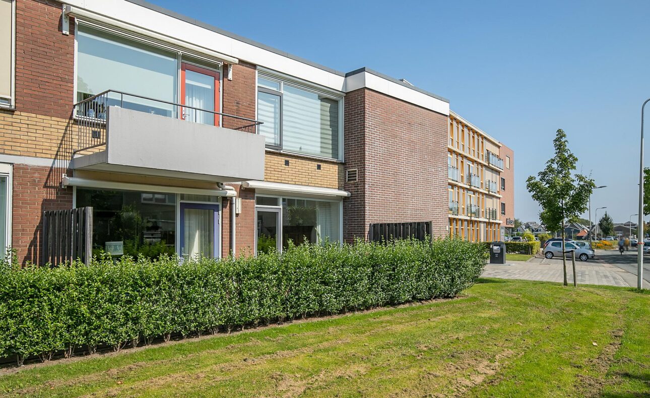Appartementencomplex met bakstenen gevels en balkons, omgeven door een groene haag en grasveld, onder een heldere blauwe lucht.