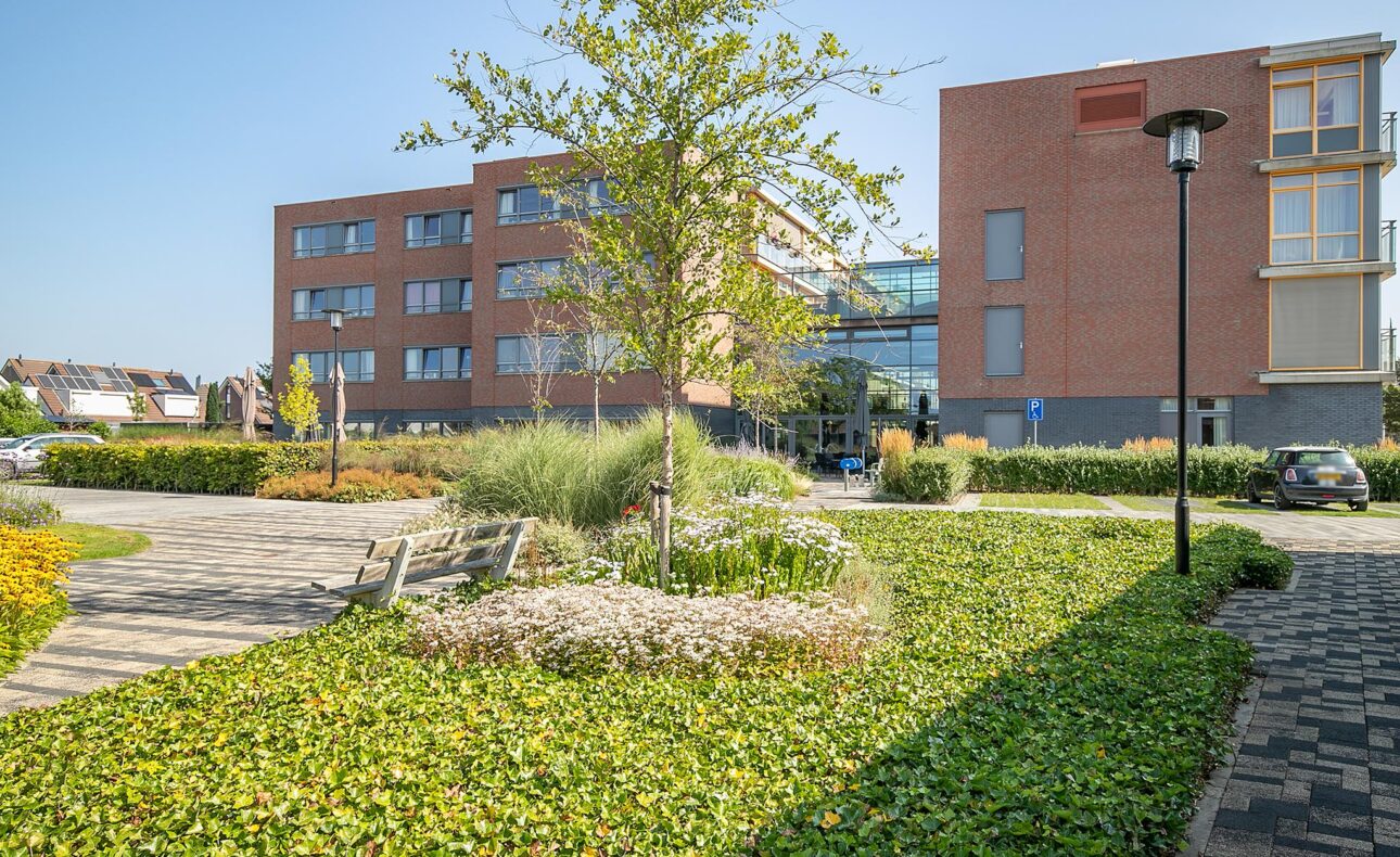 Buitenaanzicht van een modern gebouw met rode bakstenen, omgeven door groen en een geplaveid pad onder een heldere blauwe lucht.