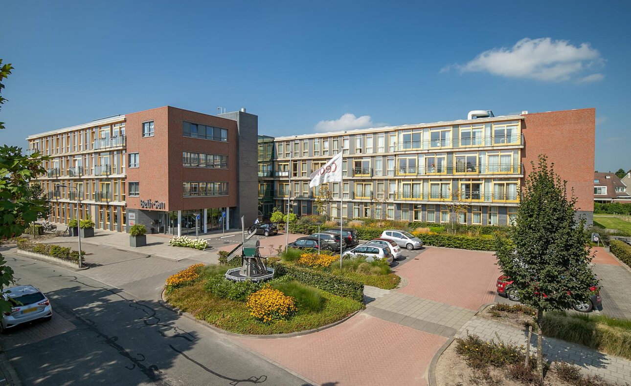 Afbeelding van een modern gebouw met meerdere verdiepingen, omgeven door groen en geparkeerde auto's op een zonnige dag.