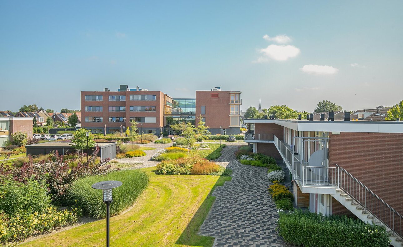 Luchtfoto van een modern zorgcentrum met groene tuinen, paden en meerdere gebouwen onder een heldere blauwe hemel.