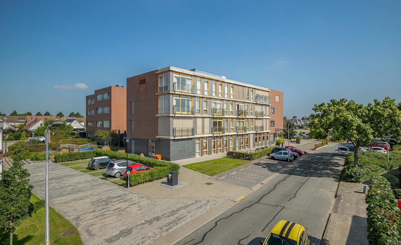 Appartementencomplex met drie verdiepingen, omgeven door parkeerplaatsen en groen, onder een heldere blauwe lucht.