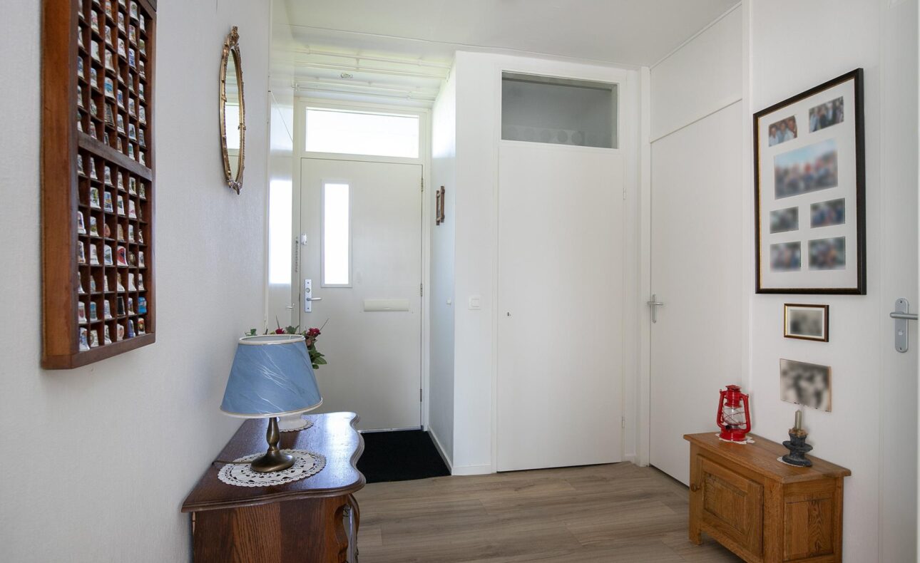 Ingang van een huis met houten vloer, een kleine tafel met een blauwe lamp, en diverse ingelijste foto's aan de witte muren.