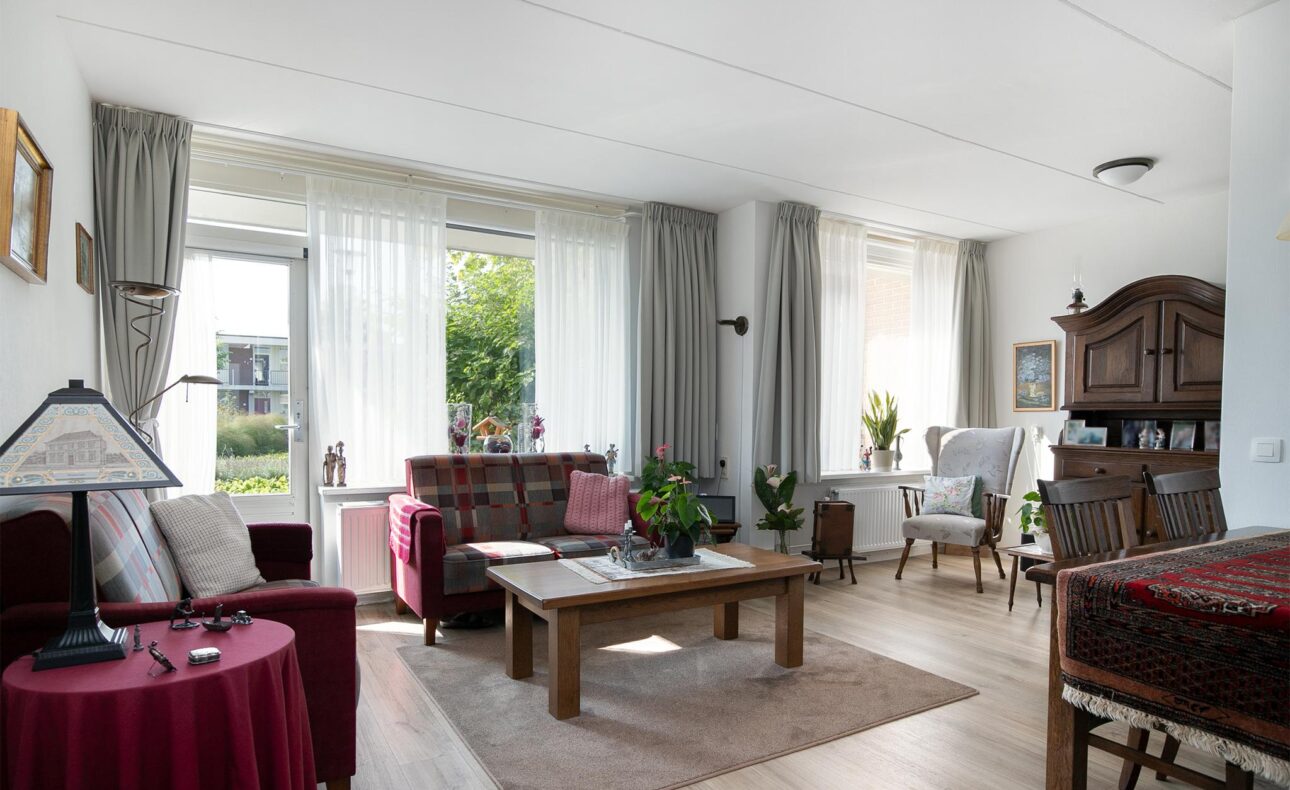 Woonkamer met houten meubels, planten en grote ramen met gordijnen. Er is een bank, fauteuils en een houten kast.