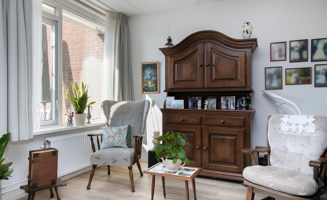 Woonkamer met houten kast, twee fauteuils, planten en schilderijen aan de muur. Groot raam met gordijnen aan de linkerkant.