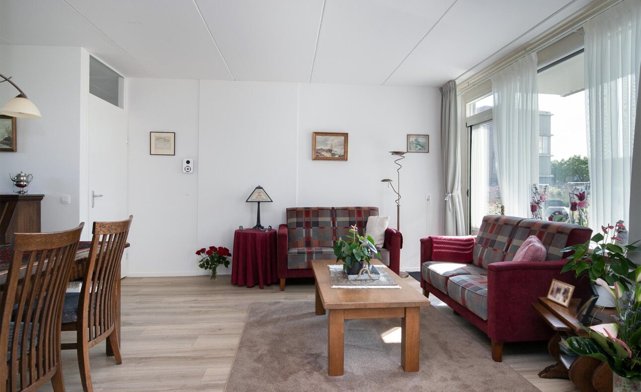 Woonkamer met houten vloer, rode banken, houten tafel en stoelen. Grote ramen met gordijnen laten veel licht binnen.