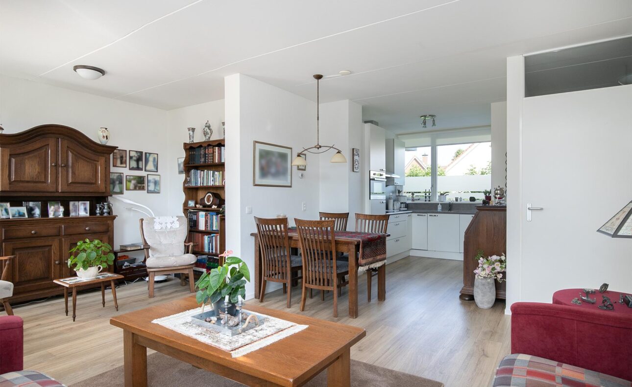 Woonkamer met houten meubels, een eettafel met stoelen, een kast, planten en een open keuken met veel lichtinval.