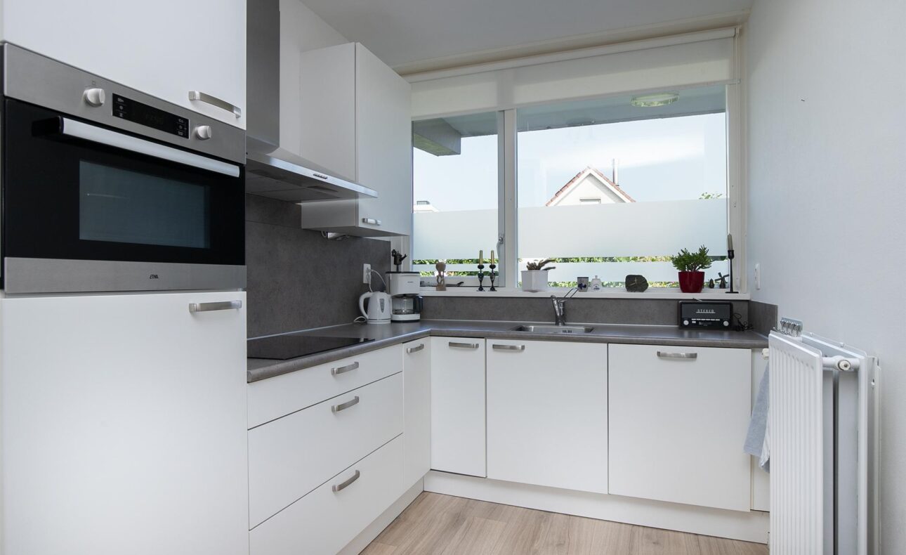 Moderne witte keuken met hoekopstelling, voorzien van inbouwapparatuur, grijs aanrechtblad en groot raam met matglas.