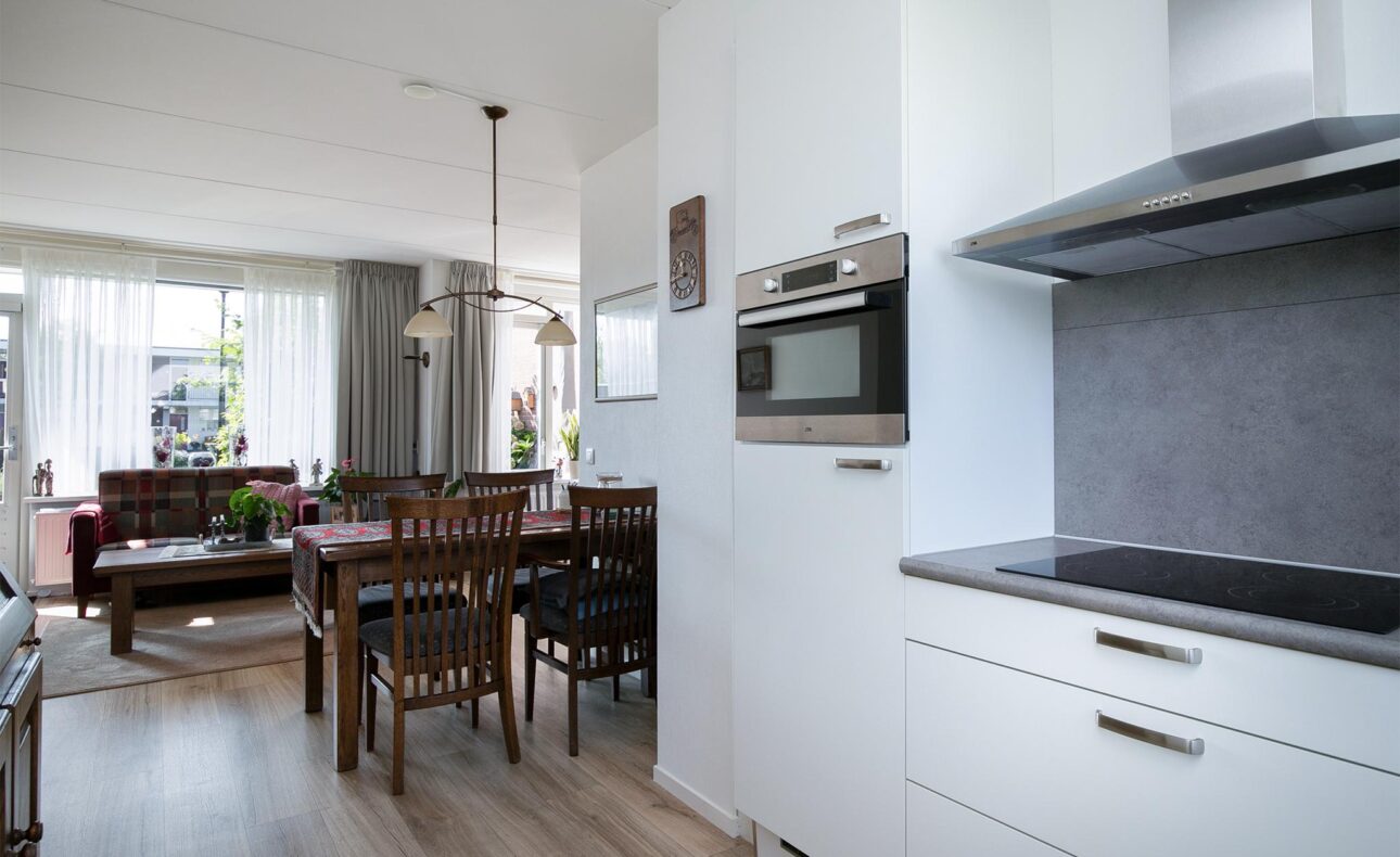 Moderne keuken met witte kasten en grijze achterwand. Op de achtergrond een eet- en woonkamer met houten meubels.