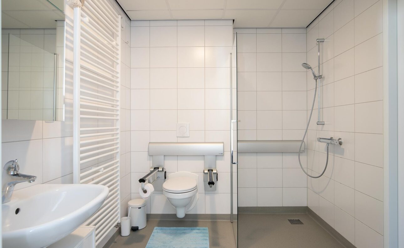 Moderne, rolstoeltoegankelijke badkamer met witte tegels, inloopdouche, toilet met beugels, wastafel en handdoekradiator.