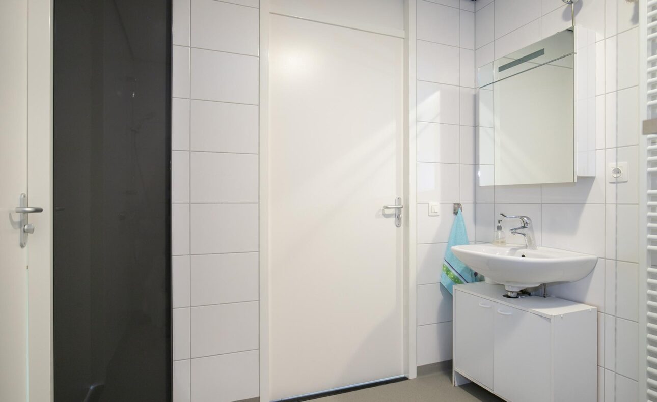 Een moderne badkamer met witte tegels, een wastafel met spiegel, en een handdoek aan de muur. Deur in het midden.