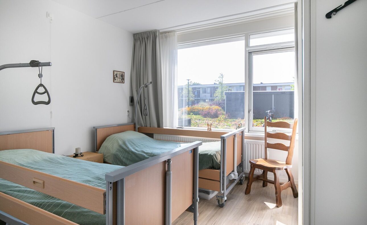 Twee ziekenhuisbedden in een lichte kamer met een houten stoel bij het raam. Er hangt een grijpstang aan het plafond.