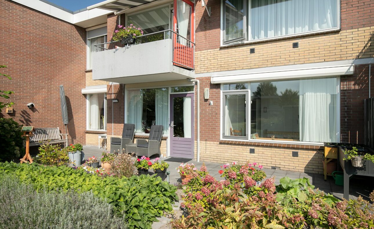 Tuin met kleurrijke bloemen en planten voor een bakstenen gebouw met balkon en grote ramen op een zonnige dag.