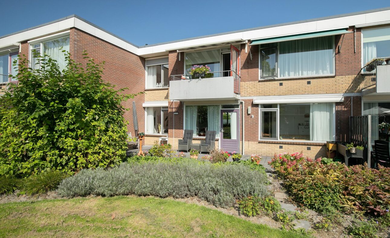 Twee verdiepingen tellend bakstenen gebouw met balkons, omgeven door groene struiken en een goed onderhouden tuin.