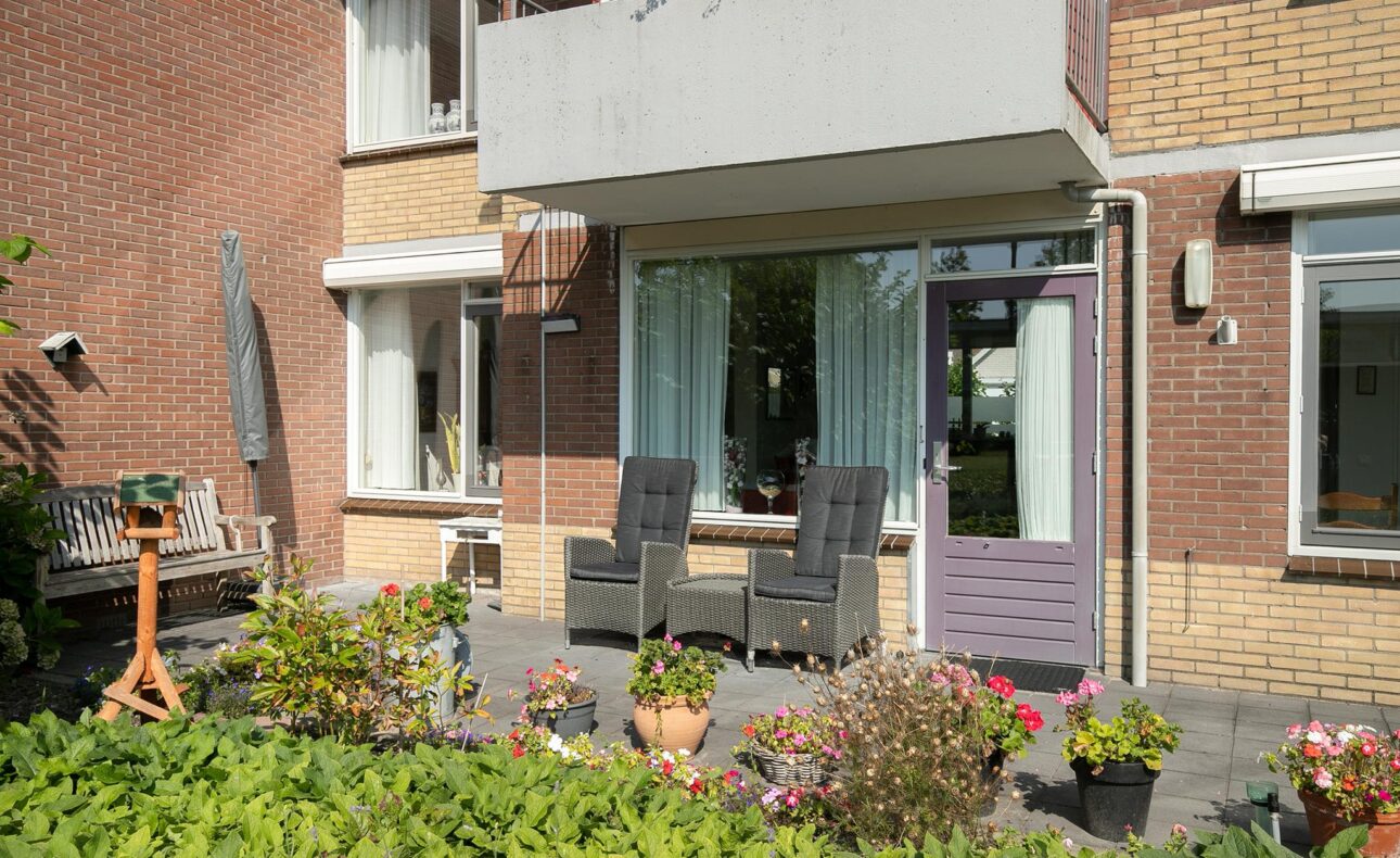 Twee stoelen en een tafel met planten op een terras voor een bakstenen huis met grote ramen en een balkon erboven.