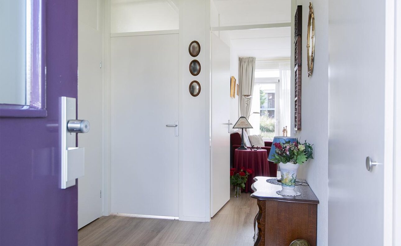 Open deur naar een lichte gang met houten vloer, een tafel met bloemen en decoraties aan de muur. Zicht op woonkamer.
