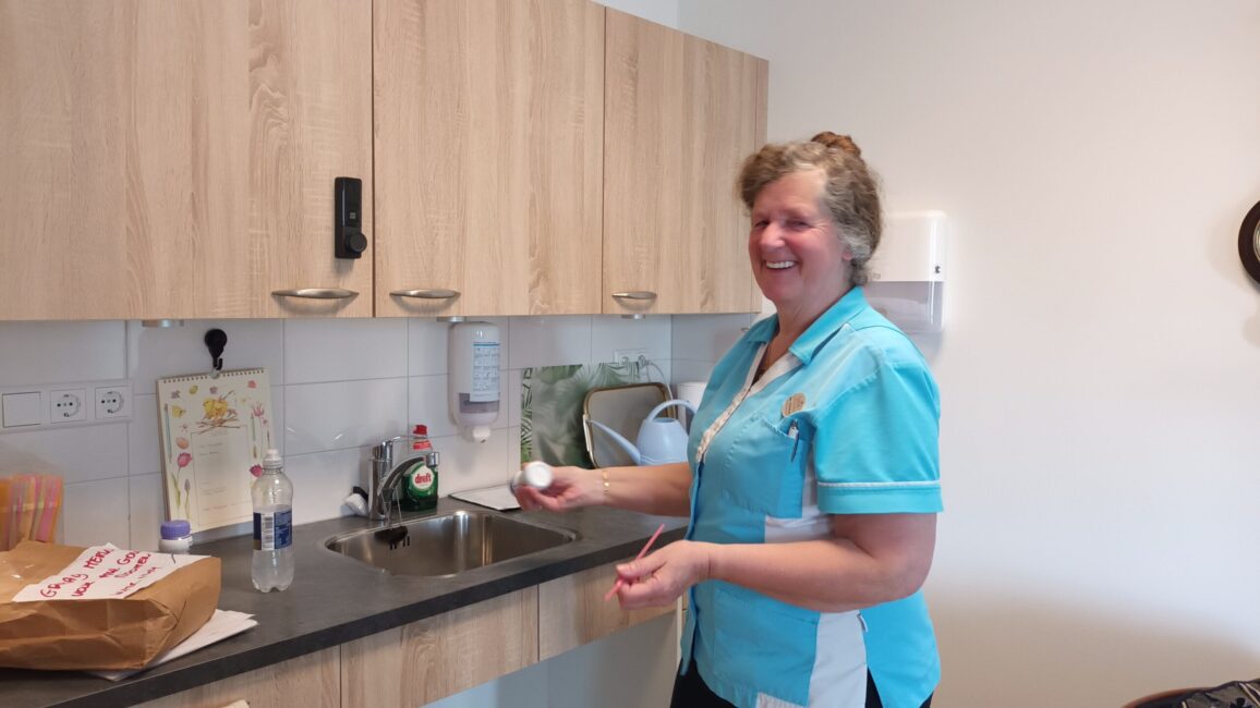 Een vrouw in een blauw uniform staat glimlachend in een keuken en houdt een ei vast. Op de achtergrond zijn keukenkastjes.