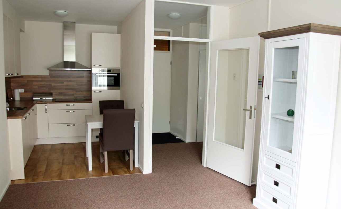 Afbeelding van een moderne keuken en eetruimte met witte kasten, een houten vloer en een eettafel met vier stoelen.