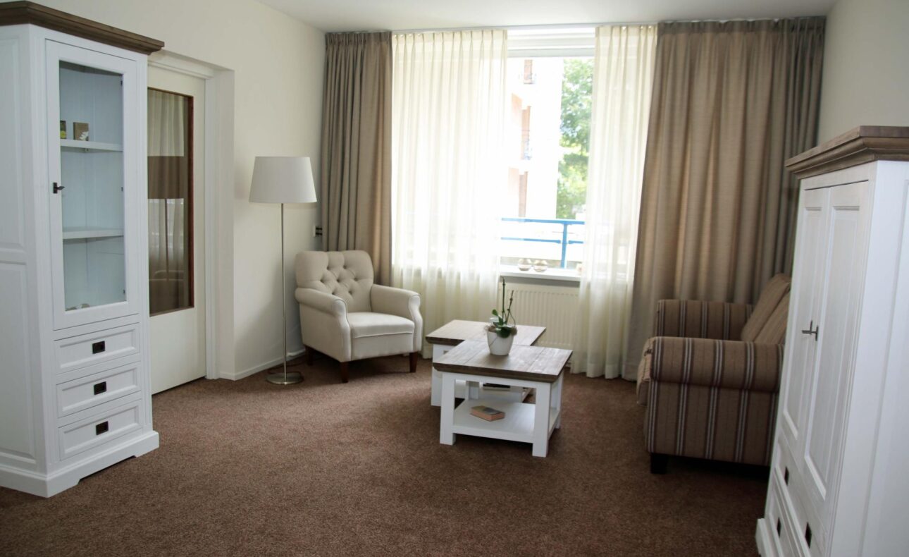 Woonkamer met witte meubels, een beige fauteuil, een gestreepte bank, een salontafel en een raam met lichte gordijnen.