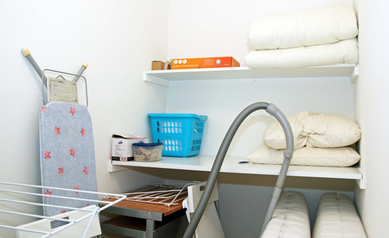 Een opbergruimte met een strijkplank, stofzuiger, wasmand, en opgevouwen dekens op planken tegen een witte muur.
