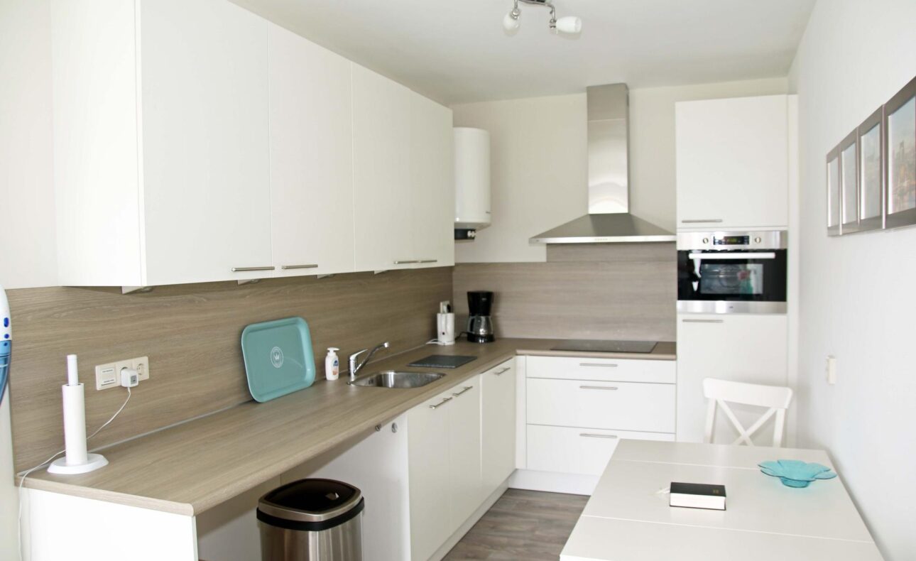 Moderne witte keuken met houten aanrechtblad, ingebouwde oven, afzuigkap en eettafel met twee stoelen. Helder verlicht.