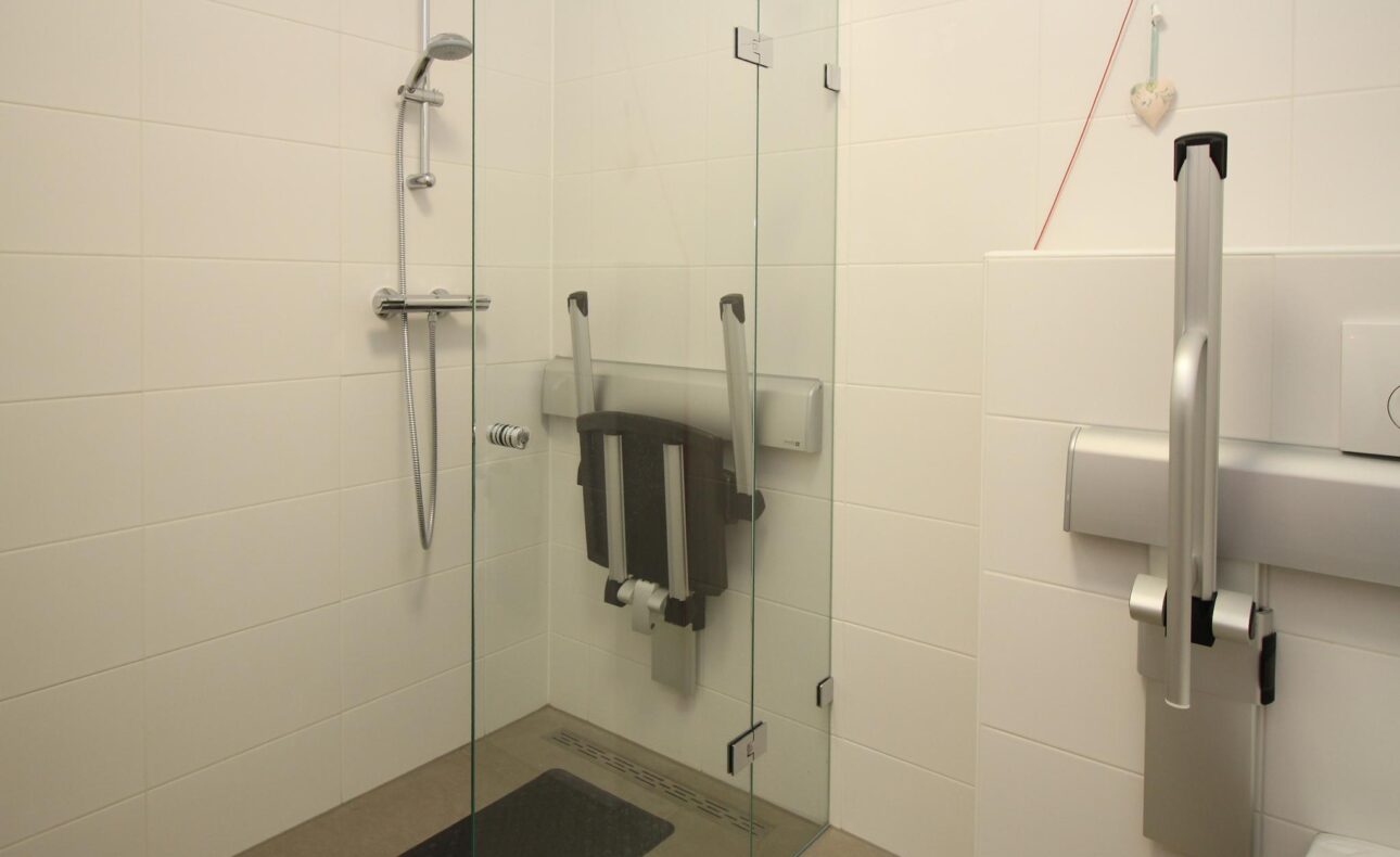 Moderne, rolstoeltoegankelijke douche met glazen wanden, een opklapbare stoel en een handgreep voor extra ondersteuning.
