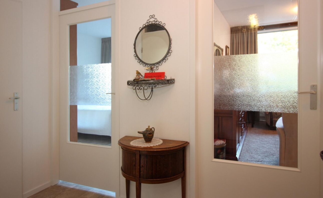 Een hal met twee deuren, een ronde spiegel aan de muur, een plank met decoraties en een klein houten tafeltje.
