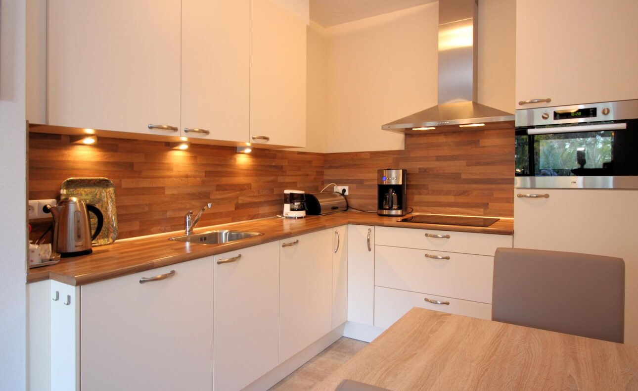 Moderne keuken met witte kasten, houten werkblad, ingebouwde oven, magnetron, en roestvrijstalen afzuigkap. Verlichte achterwand.
