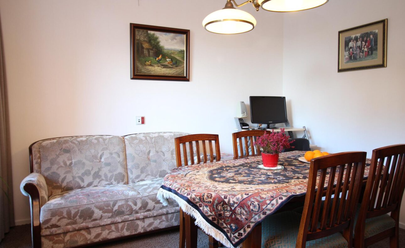 Woonkamer met een bank, een eettafel met zes stoelen, een computer op een bureau en schilderijen aan de muur.