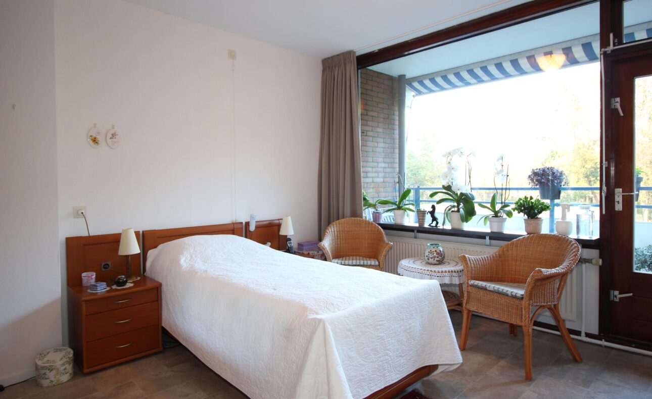 Een lichte slaapkamer met een eenpersoonsbed, twee rieten stoelen, een tafel en een raam met uitzicht op een balkon met planten.