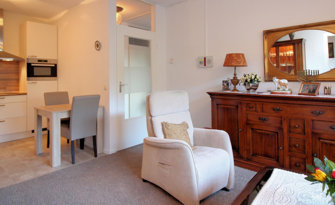 Woonkamer met een witte fauteuil, houten dressoir met spiegel, eettafel met stoelen en een open keuken op de achtergrond.