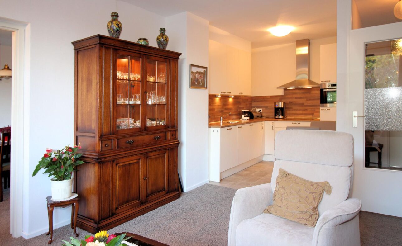 Woonkamer met houten kast, witte fauteuil, en open keuken. Planten en bloemen decoreren de ruimte. Gezellige sfeer.