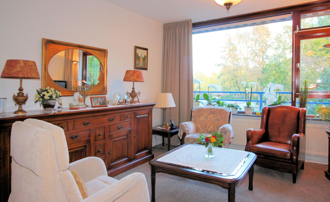 Woonkamer met houten dressoir, twee fauteuils, een salontafel en een groot raam met uitzicht op bomen en een balkon.