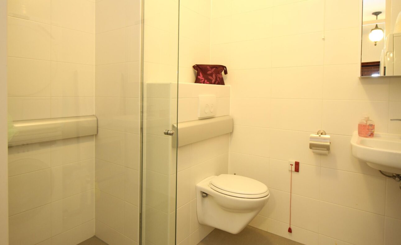 Een moderne, witte badkamer met een zwevend toilet, glazen douchewand, wastafel en handgrepen voor toegankelijkheid.