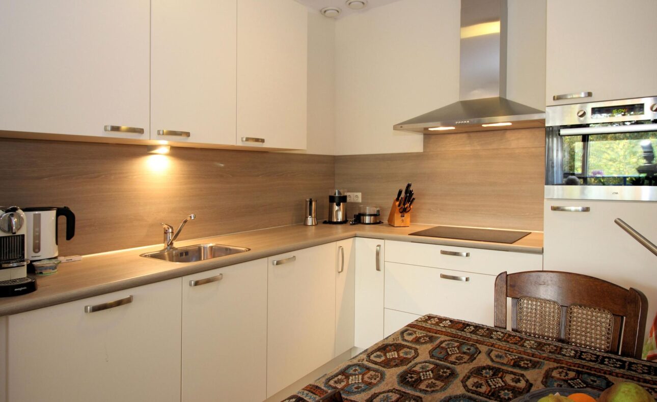 Moderne keuken met witte kasten, een fornuis, afzuigkap en een tafel met een kleurrijk tafelkleed en een fruitschaal.