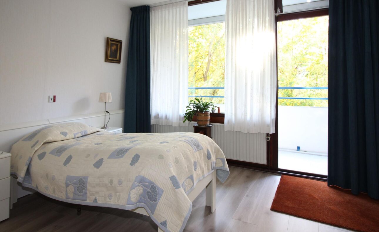 Een lichte slaapkamer met een eenpersoonsbed, witte muren, een raam met gordijnen en een deur naar een balkon met uitzicht.