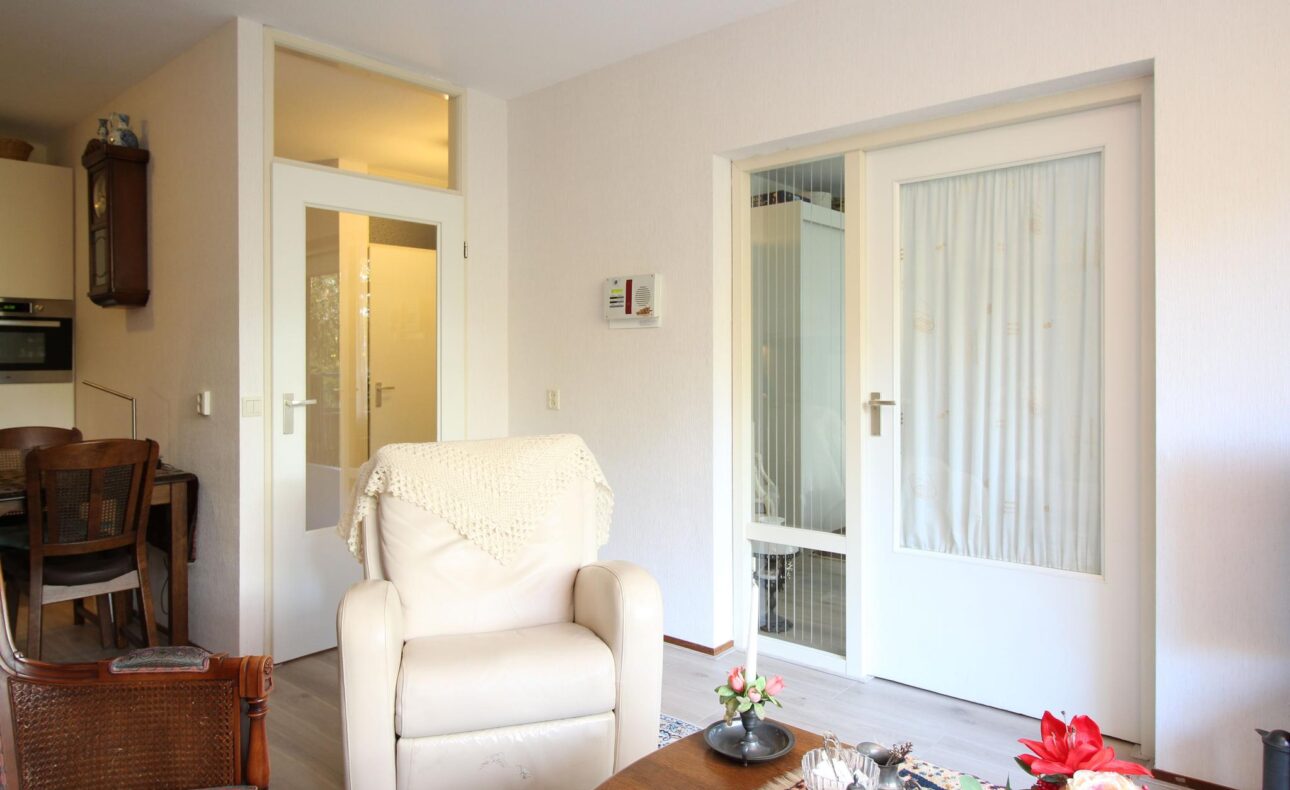 Woonkamer met een comfortabele beige stoel, houten meubels en glazen deuren. De ruimte is licht en huiselijk ingericht.