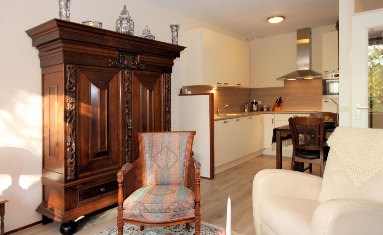 Woonkamer met een antieke houten kast, een kleurrijke stoel, een beige bank en een moderne open keuken op de achtergrond.