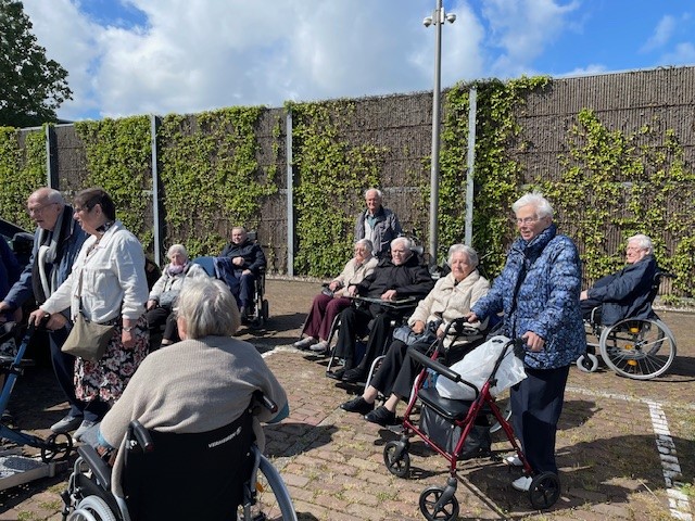 Groep ouderen geniet van een zonnige dag buiten, sommigen in rolstoelen en anderen met rollators, voor een groene muur.