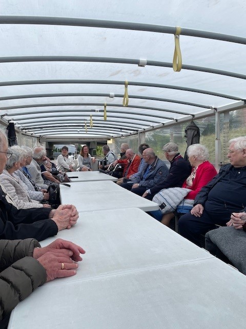 Groep ouderen zit aan een lange tafel in een overdekte ruimte, gezellig pratend tijdens een uitje. Tafels zijn wit gedekt.
