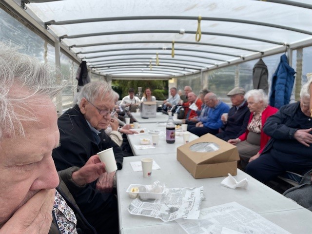 Groep ouderen zit aan een lange tafel in een overdekte ruimte, genietend van een gezamenlijke maaltijd en elkaars gezelschap.