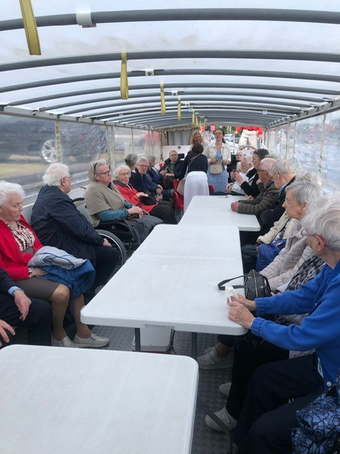 Groep ouderen zit aan lange tafels in een overdekte boot tijdens een uitje. Ze lijken te genieten van de gezamenlijke activiteit.