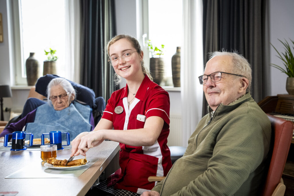 Een zorgmedewerker in rood uniform zit glimlachend aan tafel met twee oudere mensen in een gezellige kameromgeving.