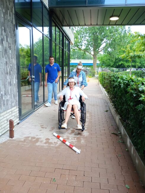 Een vrouw in een rolstoel wordt buiten geduwd door een andere vrouw. Een man loopt naast hen. Ze lachen en genieten van de dag.