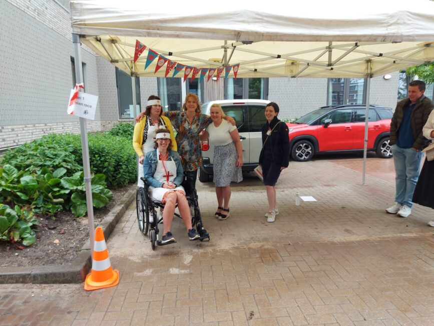 Groep mensen poseert onder een partytent met vlaggetjes. Een persoon zit in een rolstoel. Er staat een rode auto op de achtergrond.
