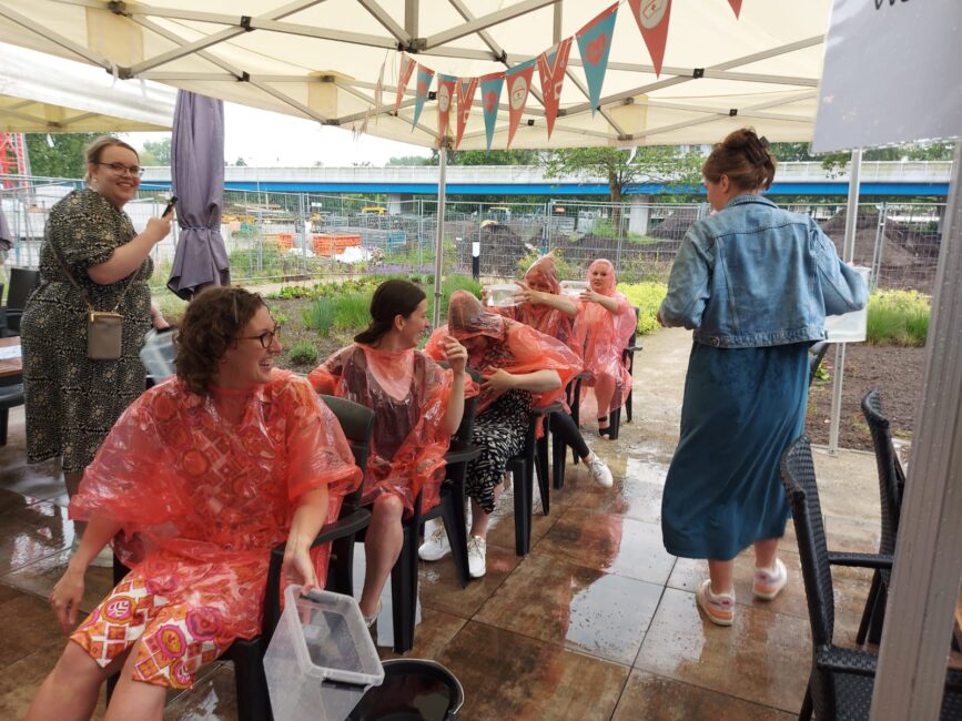 Mensen in regenponcho's zitten op stoelen onder een tent, terwijl een vrouw in een blauwe jas langsloopt. Het regent buiten.